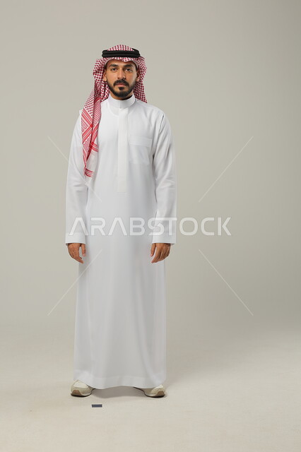 الاهتمام بالمظهر الخارجي، الوقوف باستقامة وانضباط، مفهوم الرجولة والأناقه، الثقة بالنفس والشعور بالقوة، بورتريه لشاب عربي خليجي سعودي يرتدي الشماغ والثوب التقليدي يضع يديه على جانبه و ينظر إلى الكاميرا بإيماءات السرور، صورة الجسم بالطول الكامل، خلفية رمادية
