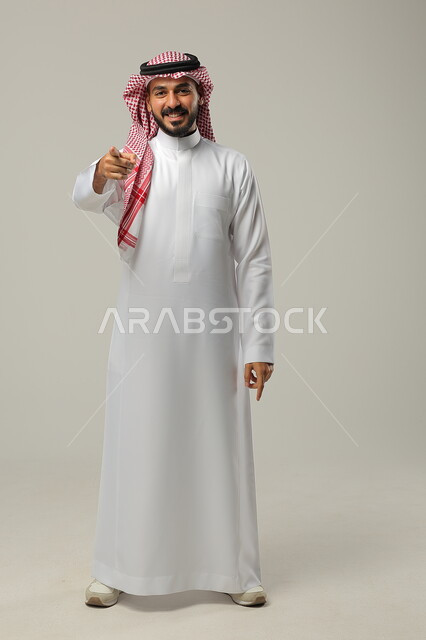 ايماءات التأكيد وتحديد الشيء المقابل, الإشارة الى شيء ما بحركات التواجد هنا, بورتريه لرجل عربي خليجي سعودي يرتدي الثوب التقليدي والشماغ يرفع يديه للأمام بتعابير السرور, صورة الجسم بالطول الكامل, خلفية رمادية
