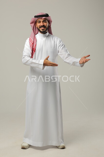 الإشارة الى شيء ما بحركات التواجد هنا, ايماءات التأكيد وتحديد الشيء المقابل, بورتريه لرجل عربي خليجي سعودي يرتدي الثوب التقليدي والشماغ يرفع يديه بتعابير الدلالة, صورة الجسم بالطول الكامل, خلفية رمادية