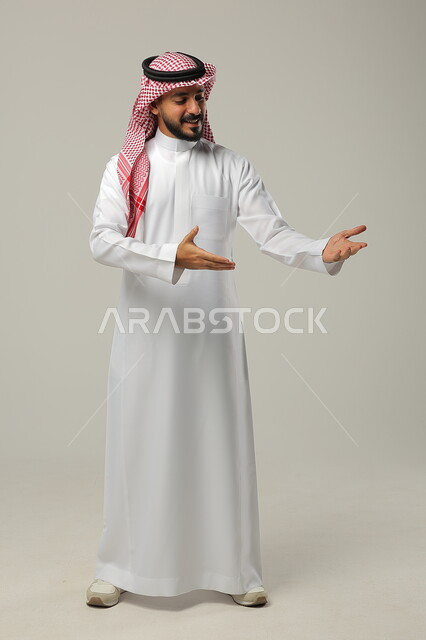 Pointing to something with gestures of being here, gestures of affirmation and identification of the opposite, portrait of a Saudi Arabian Gulf man wearing traditional thobe and shemagh raising his hands with expressions of significance, full-length body image, gray background