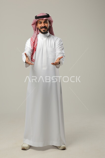 الإشارة الى شيء ما بحركات التواجد هنا, ايماءات التأكيد وتحديد الشيء المقابل, بورتريه لرجل عربي خليجي سعودي يرتدي الثوب التقليدي والشماغ يرفع يديه بتعابير الدلالة, صورة الجسم بالطول الكامل, خلفية رمادية