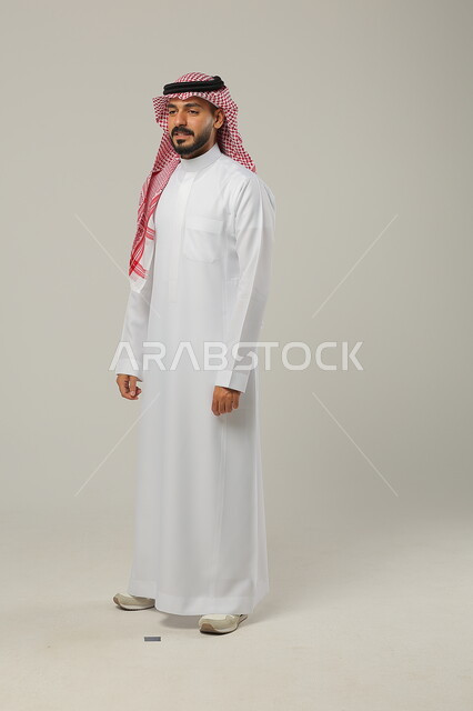  مفهوم الرجولة والأناقه، الوقوف باستقامة وانضباط، الاهتمام بالمظهر الخارجي، الثقة بالنفس والشعور بالقوة، بورتريه لشاب عربي خليجي سعودي مبتسم يرتدي الشماغ والثوب التقليدي يضع يديه على جانبه و ينظر إلى الكاميرا بإيماءات السرور، صورة الجسم بالطول الكامل، خلفية رمادية