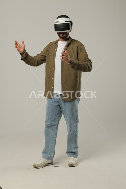 التفاعل والاندماج مع ألعاب الواقع الافتراضي, تقنيات وأجهزة متطورة, محاكاة العالم الخيالي ومحاولة لمس عنصر ما, إيماءات الدهشة والاستمتاع, بورتريه لرجل عربي سعودي خليجي يرتدي الزي الكاجوال ونظارة في آر ثلاثية الأبعاد, صورة الجسم بالطول الكامل, خلفية رمادية
