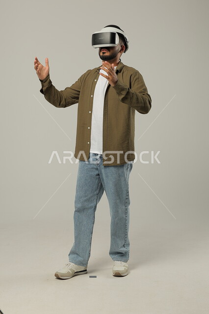Simulating the imaginary world and trying to touch an item, gestures of surprise and enjoyment, portrait of a Saudi Arabian Gulf man wearing casual clothes and 3D VR glasses, interaction and integration with virtual reality games, advanced technologies and devices, full-length body image, gray background