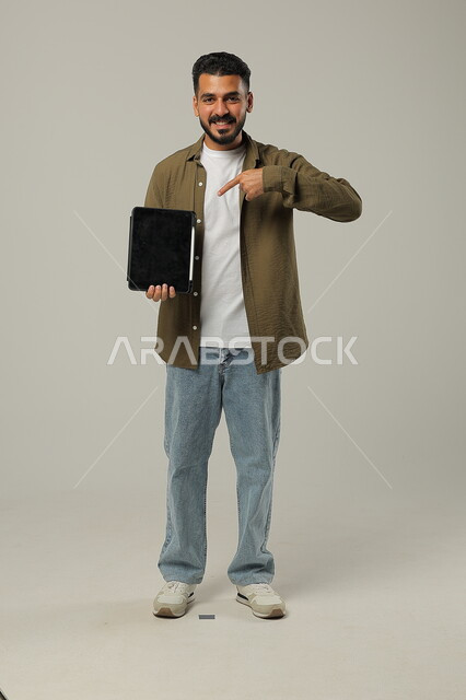 الاشارة الى جهاز التابلت والنظر الى الكاميرا بتعابير وايماءات الاعجاب، بورتريه لشاب عربي خليجي سعودي مبتسم يرتدي الزي الكاجوال يقوم باستخدام التقنيات الحديثه المتطورة في الحياة العملية عبر الأجهزة اللوحية، صورة الجسم بالطول الكامل، خلفية رمادية