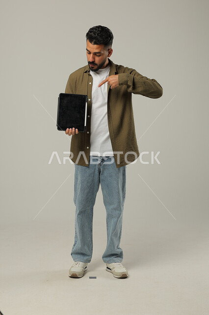 الاشارة الى جهازالتابلت والنظر اليه بتعابير وايماءات الاعجاب، بورتريه لشاب عربي خليجي سعودي مبتسم يرتدي الزي الكاجوال يقوم باستخدام التقنيات الحديثه المتطورة في الحياة العملية عبر الأجهزة اللوحية، صورة الجسم بالطول الكامل، خلفية رمادية