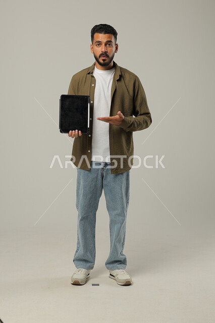 النظر الى الكاميرا والاشارة الى جهازالتابلت بتعابير وايماءات الاعجاب، بورتريه لشاب عربي خليجي سعودي مبتسم يرتدي الزي الكاجوال يقوم باستخدام التقنيات الحديثه المتطورة في الحياة العملية عبر الأجهزة اللوحية، صورة الجسم بالطول الكامل، خلفية رمادية