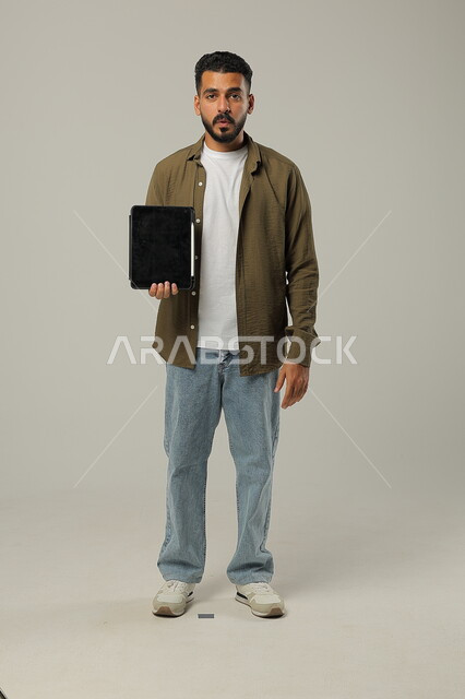 استخدام التقنيات الحديثه المتطورة في الحياة العملية عبر الأجهزة اللوحية، النظر الى الكاميرا بتعابير وايماءات الاعجاب، بورتريه لشاب عربي خليجي سعودي مبتسم يرتدي الزي الكاجوال يحمل بيده جهازالتابلت، صورة الجسم بالطول الكامل، خلفية رمادية