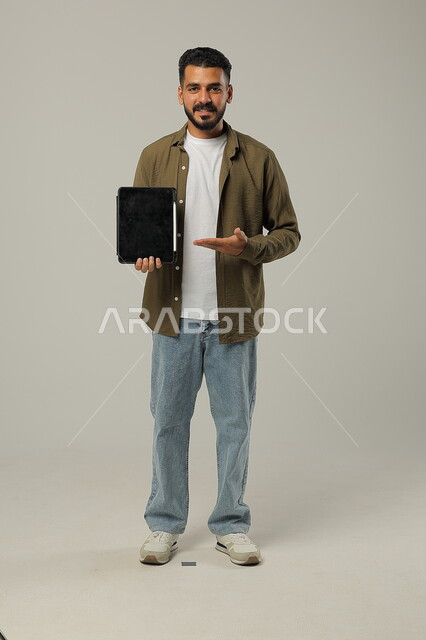 النظر الى الكاميرا والاشارة الى جهازالتابلت بتعابير وايماءات الاعجاب، بورتريه لشاب عربي خليجي سعودي مبتسم يرتدي الزي الكاجوال يقوم باستخدام التقنيات الحديثه المتطورة في الحياة العملية عبر الأجهزة اللوحية، صورة الجسم بالطول الكامل، خلفية رمادية