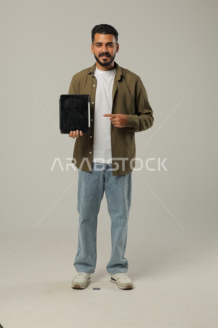الاشارة الى جهاز التابلت والنظر الى الكاميرا بتعابير وايماءات الاعجاب، بورتريه لشاب عربي خليجي سعودي مبتسم يرتدي الزي الكاجوال يقوم باستخدام التقنيات الحديثه المتطورة في الحياة العملية عبر الأجهزة اللوحية، صورة الجسم بالطول الكامل، خلفية رمادية