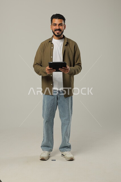 Expressions and gestures of admiration using modern advanced technologies in practical life via tablets, portrait of a smiling Saudi Arabian Gulf young man wearing casual clothes working on a tablet looking at the camera, full body image, gray background