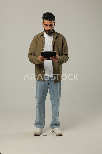 استخدام التقنيات الحديثه المتطورة في الحياة العملية عبر الأجهزة اللوحية، تعابير وايماءات الاندماج، بورتريه لشاب عربي خليجي سعودي مبتسم يرتدي الزي الكاجوال يقوم بالعمل على جهازالتابلت، صورة الجسم بالطول الكامل، خلفية رمادية