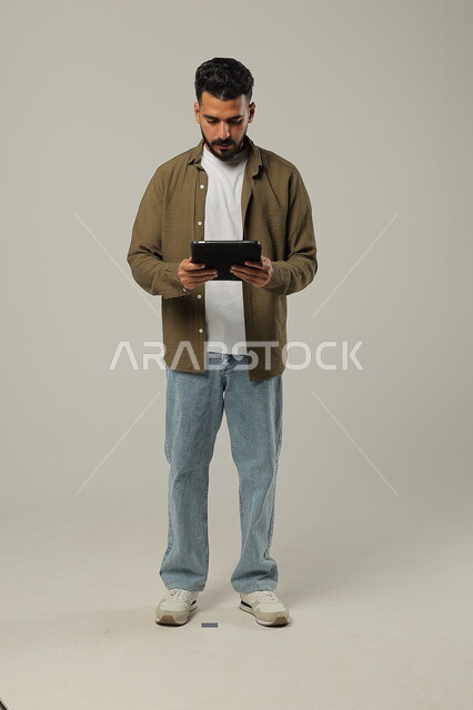 Using advanced modern technologies in practical life via tablets, expressions and gestures of integration, portrait of a smiling Saudi Arabian Gulf young man wearing casual clothes working on a tablet, full body image, gray background