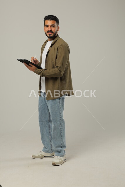 ايماءت الاعجاب باستخدام التقنيات الحديثه المتطورة في الحياة العملية عبر الأجهزة اللوحية، بورتريه لشاب عربي خليجي سعودي مبتسم يرتدي الزي الكاجوال يقوم بالعمل على جهازالتابلت، صورة الجسم بالطول الكامل، خلفية رمادية