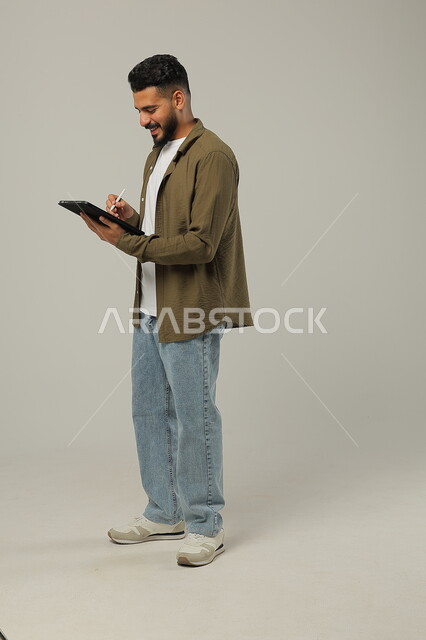 ايماءت الاعجاب باستخدام التقنيات الحديثه المتطورة في الحياة العملية عبر الأجهزة اللوحية، بورتريه لشاب عربي خليجي سعودي مبتسم يرتدي الزي الكاجوال يقوم بالعمل على جهازالتابلت، صورة الجسم بالطول الكامل، خلفية رمادية