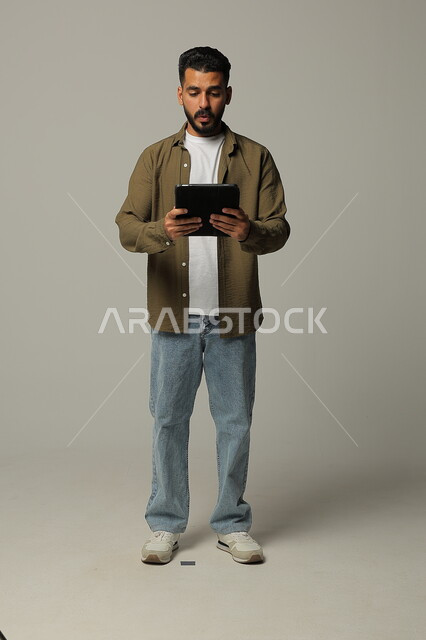 Expressions and gestures of integration, using modern advanced technologies in practical life via tablets, portrait of a smiling young Saudi Arabian Gulf man wearing casual clothes working on a tablet, full body image, gray background