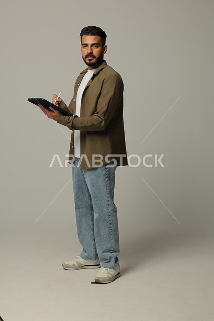 العمل على جهازالتابلت، بورتريه لشاب عربي خليجي سعودي مبتسم يرتدي الزي الكاجوال ينظر الى الكاميرا بايماءت الاعجاب باستخدام التقنيات الحديثه المتطورة في الحياة العملية عبر الأجهزة اللوحية، صورة الجسم بالطول الكامل، خلفية رمادية