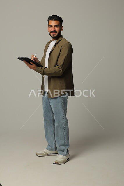 Working on tablet, portrait of a smiling Saudi Arabian Gulf young man in casual clothes looking at the camera with gestures of admiration using modern advanced technologies in practical life via tablets, full body image, gray background