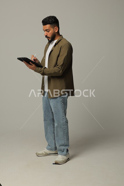 Using modern advanced technologies in practical life via tablets, expressions and gestures of integration, portrait of a smiling Saudi Arabian Gulf young man wearing casual clothes working on a tablet, full body image, gray background