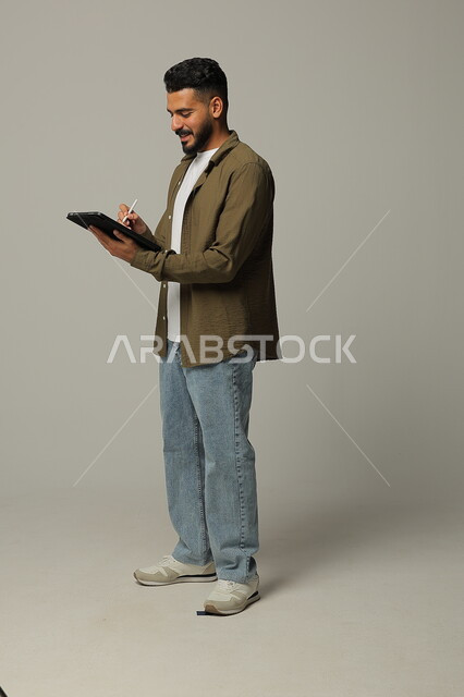 Using modern advanced technologies in practical life via tablets, expressions and gestures of integration, portrait of a smiling Saudi Arabian Gulf young man wearing casual clothes working on a tablet, full body image, gray background