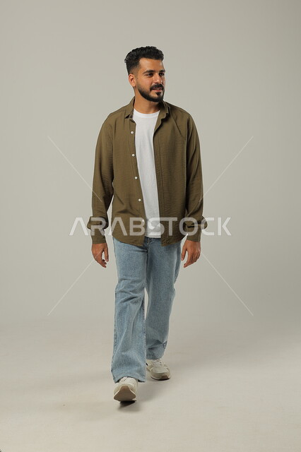 المشي والذهاب الى مكان ما، مفهوم الاهتمام بالمظهر الخارجي والأناقة، بورتريه لرجل عربي خليجي سعودي مبتسم يرتدي الزي الكاجوال ينظر إلى شيء ما بإيماءات البشاشة والسرور، صورة الجسم بالطول الكامل، خلفية رمادية