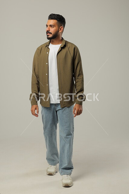 المشي والذهاب الى مكان ما، مفهوم الاهتمام بالمظهر الخارجي والأناقة، بورتريه لرجل عربي خليجي سعودي مبتسم يرتدي الزي الكاجوال ينظر إلى شيء ما بإيماءات البشاشة والسرور، صورة الجسم بالطول الكامل، خلفية رمادية