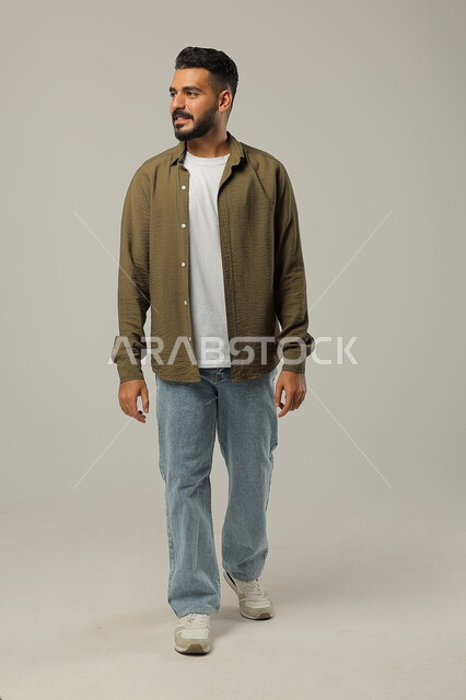 Walking and going somewhere, concept of caring about appearance and elegance, portrait of a smiling Saudi Arabian Gulf man in casual clothes looking at something with gestures of cheerfulness and pleasure, full body image, gray background