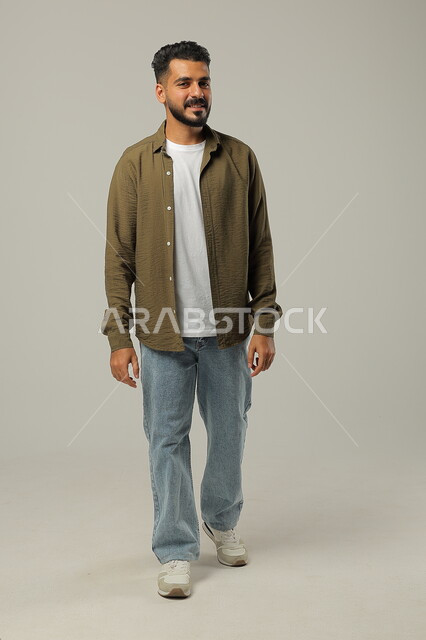 المشي والذهاب الى مكان ما، مفهوم الاهتمام بالمظهر الخارجي والأناقة، بورتريه لرجل عربي خليجي سعودي مبتسم يرتدي الزي الكاجوال ينظر إلى شيء ما بإيماءات البشاشة والسرور، صورة الجسم بالطول الكامل، خلفية رمادية