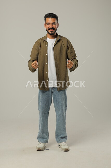 حركات تدل على الكسب والربح وانجاز المهام، الشغف في تشجيع فريق كرة القدم، ايماءات الفوز والانتصار وتحقيق الأهداف، بورتريه لشاب عربي خليجي سعودي يرتدي الزي الكاجوال يرفع يديه بتفاعل وحماس، صورة الجسم بالطول الكامل، خلفية رمادية