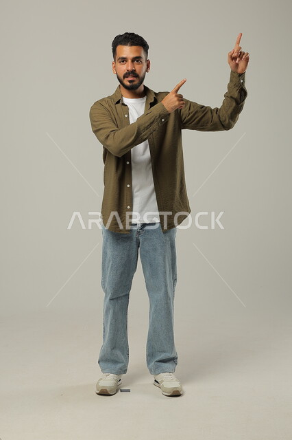 Standing straight and looking at camera, gestures of inquiry and pointing at something, portrait of smiling Saudi Arabian Gulf man in casual wear raising finger up with clarifying gestures, full body portrait, gray background