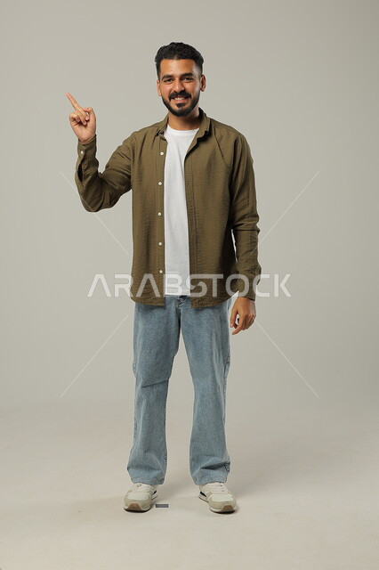 Standing straight and looking at camera, gestures of inquiry and pointing at something, portrait of smiling Saudi Arabian Gulf man in casual wear raising finger up with clarifying gestures, full body portrait, gray background