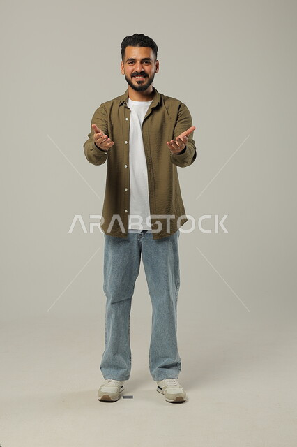 الوقوف باستقامة والنظر إلى الكاميرا، الترحيب والاستقبال بحب وبشاشة، بورتريه لشاب عربي سعودي خليجي يرتدي الزي الكاجوال يفتح كفيه بإيماءات الفرح والسرور, صورة الجسم بالطول الكامل, خلفية رمادية