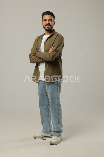 الوقوف بأيدي متقاطعه بتعابير الثقة بالنفس، بورتريه لشاب وسيم عربي خليجي سعودي يرتدي الزي الكاجوال يقف باستقامة وثقه بالنفس، مفهوم الاناقه و الاهتمام بالمظهر الخارجي، صورة الجسم بالطول الكامل، خلفية رمادية