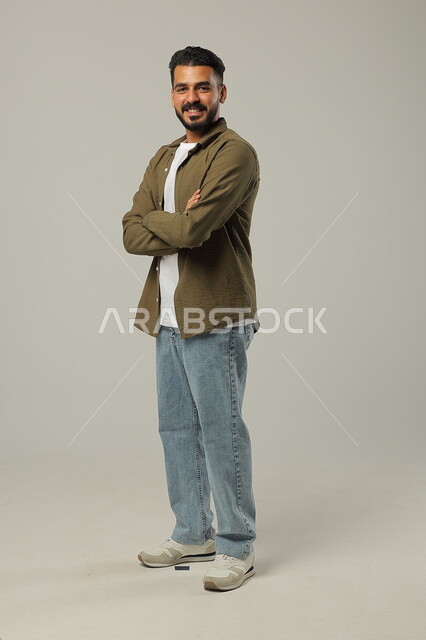الوقوف بأيدي متقاطعه بتعابير الثقة بالنفس، بورتريه لشاب وسيم عربي خليجي سعودي يرتدي الزي الكاجوال يقف باستقامة وثقه بالنفس، مفهوم الاناقه و الاهتمام بالمظهر الخارجي، صورة الجسم بالطول الكامل، خلفية رمادية