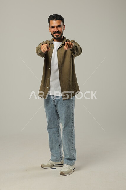 Gestures of inquiry and pointing to something, standing straight and looking at camera, portrait of smiling Saudi Arabian Gulf man in casual wear raising finger up with clarifying gestures, full body photo, gray background