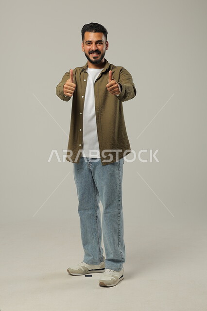 الشعور بالإعجاب والرضا والسعاده، النظر إلى الكاميرا بايماءات القبول والموافقه, بورتريه لشاب وسيم عربي خليجي سعودي مبتسم يرتدي الزي الكاجوال ويرفع أصبع الابهام بعلامة الجودة، صورة الجسم بالطول الكامل، خلفية رمادية