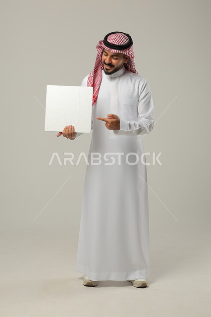 Using illustrations and educational tools, advertisements and marketing presentations, looking at a white square mockup, portrait of a smiling young Saudi Arabian Gulf man wearing a traditional shemagh and thobe holding a blank white square board and pointing to it, full-length body image, gray background