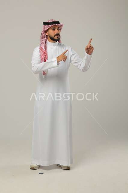 الإشارة الى شيئ ما بحركات التواجد هنا, ايماءات التأكيد وتحديد الشيء المقابل, بورتريه لرجل عربي خليجي سعودي يرتدي الثوب التقليدي والشماغ يرفع يديه للأمام بتعابير السرور, صورة الجسم بالطول الكامل, خلفية رمادية