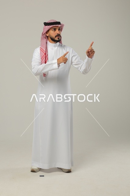 الإشارة الى شيئ ما بحركات التواجد هنا, ايماءات التأكيد وتحديد الشيء المقابل, بورتريه لرجل عربي خليجي سعودي يرتدي الثوب التقليدي والشماغ يرفع يديه للأمام بتعابير السرور, صورة الجسم بالطول الكامل, خلفية رمادية