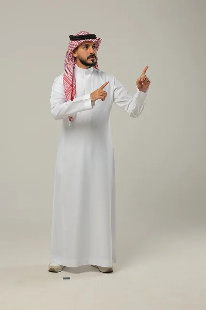 Pointing to something with gestures of being here, gestures of affirmation and identification of the opposite, portrait of a Saudi Arabian Gulf man wearing a traditional thobe and shemagh raising his hands forward with expressions of pleasure, full-length body image, gray background