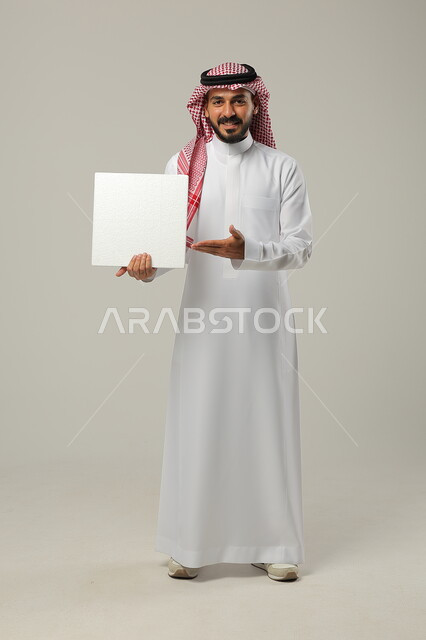 إعلانات وعروض تسويقية، موك اب مربع أبيض، استخدام لوحات توضيحية ووسائل تعليمية، بورتريه لشاب عربي خليجي سعودي مبتسم يرتدي الشماغ والثوب التقليدي ينظر إلى الكاميرا يمسك بيده لوحه بيضاء مربعة فارغة يشير اليها، صورة الجسم بالطول الكامل، خلفية رمادية