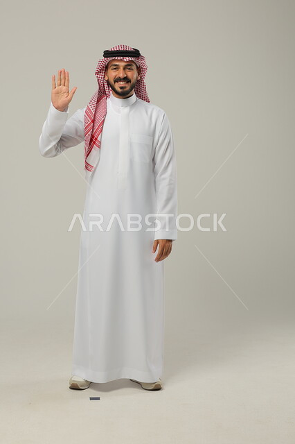 ايماءات إلقاء التحية والسلام والسعادة بالترحيب، بورتريه لرجل عربي خليجي سعودي يرتدي الثوب والشماغ التقليدي يقوم برفع يده اليمنى للأعلى، صورة الجسم بالطول الكامل، خلفية رمادية
