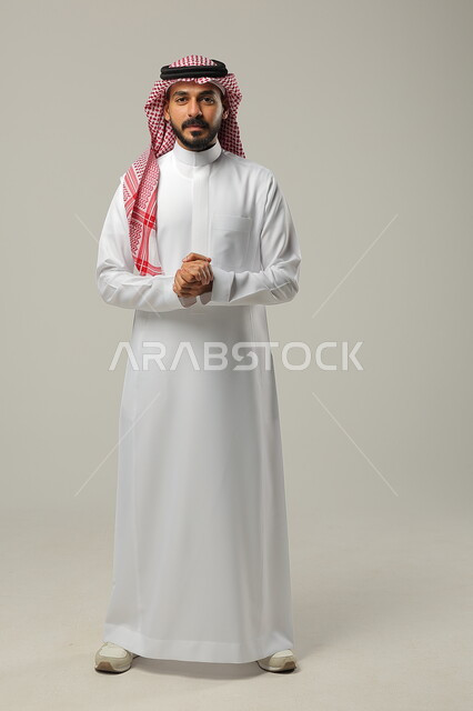 إيماءات التمعن والتركيز، إيجاد حل لمشكلة ما, التفكير و التأمل و شرود الذهن والتخطيط للمستقبل, بورتريه لشاب عربي خليجي سعودي يرتدي الثوب التقليدي والشماغ يقوم باغلاق كفيه على بعضها، صورة الجسم بالطول الكامل، خلفية رمادية