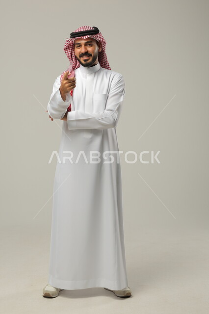 ايماءات التأكيد وتحديد الشيء المقابل, الإشارة الى شيء ما بحركات التواجد هنا, بورتريه لرجل عربي خليجي سعودي يرتدي الثوب التقليدي والشماغ يرفع يديه للأمام بتعابير السرور, صورة الجسم بالطول الكامل, خلفية رمادية