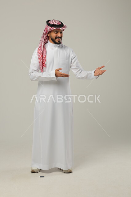Paying attention to external appearance, standing upright and disciplined, portrait of a Saudi Gulf Arab man wearing a traditional thobe and shemagh, pointing to something with his hands, full-length body portrait, gray background