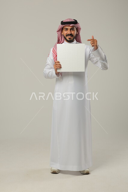 التسويق الإعلاني على اللوحات الدعائية, النظر إلى الكاميرا بإيماءات السعادة و السرور, بورتريه لرجل عربي خليجي سعودي مبتسم يرتدي الثوب التقليدي و الشماغ يشير بإصبع السبابة إلى اللوحة الفارغة, صورة للجسم بالطول الكامل, خلفية رمادية