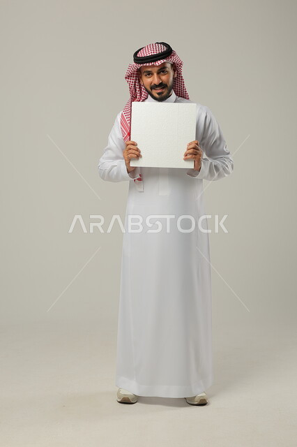 الترويج الإعلاني على موك اب أبيض, النظر إلى الكاميرا بإيماءات السعادة و السرور, بورتريه لرجل عربي خليجي سعودي يرتدي الثوب التقليدي و الشماغ يمسك بيديه لوحة فارغة, صورة للجسم بالطول الكامل, خلفية رمادية