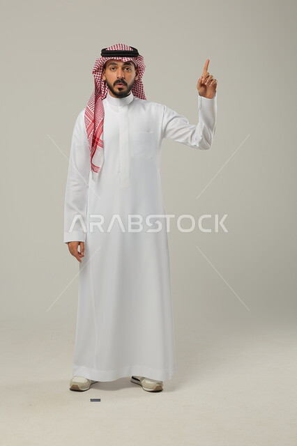 النظر إلى الكاميرا بإيماءات الدهشة و الاستغراب, الإشارة بإصبع السبابة إلى الأعلى, بورتريه لرجل عربي خليجي سعودي يرتدي الثوب التقليدي و الشماغ يشير إلى شيء ما, صورة للجسم بالطول الكامل, خلفية رمادية