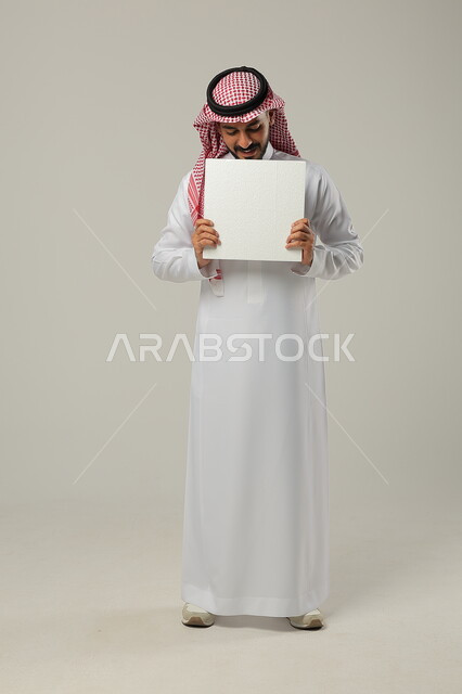 الترويج الإعلاني على موك اب أبيض, استخدام لوحات توضيحية للعروض التسويقية, بورتريه لرجل عربي خليجي سعودي يرتدي الثوب التقليدي و الشماغ يحمل بيديه لوحة بيضاء فارغة و ينظر اليها, صورة للجسم بالطول الكامل, خلفية رمادية