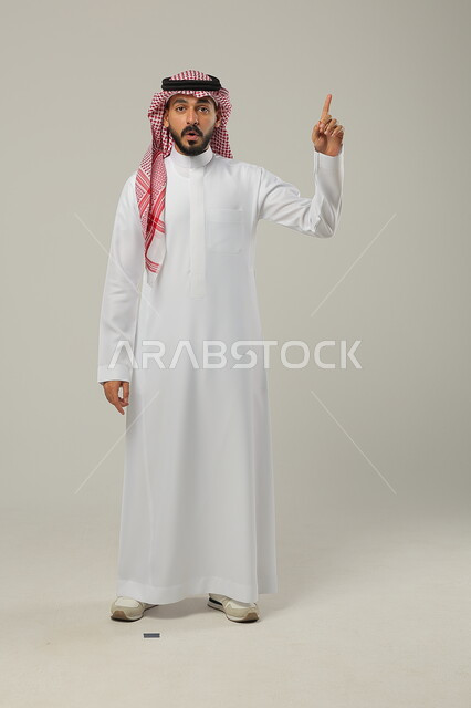 الإشارة بإصبع السبابة إلى لأعلى, النظر إلى الكاميرا بإيماءات الدهشة و الاستغراب, بورتريه لرجل عربي خليجي سعودي يرتدي الثوب التقليدي و الشماغ يشير إلى شيء ما, صورة للجسم بالطول الكامل, خلفية رمادية